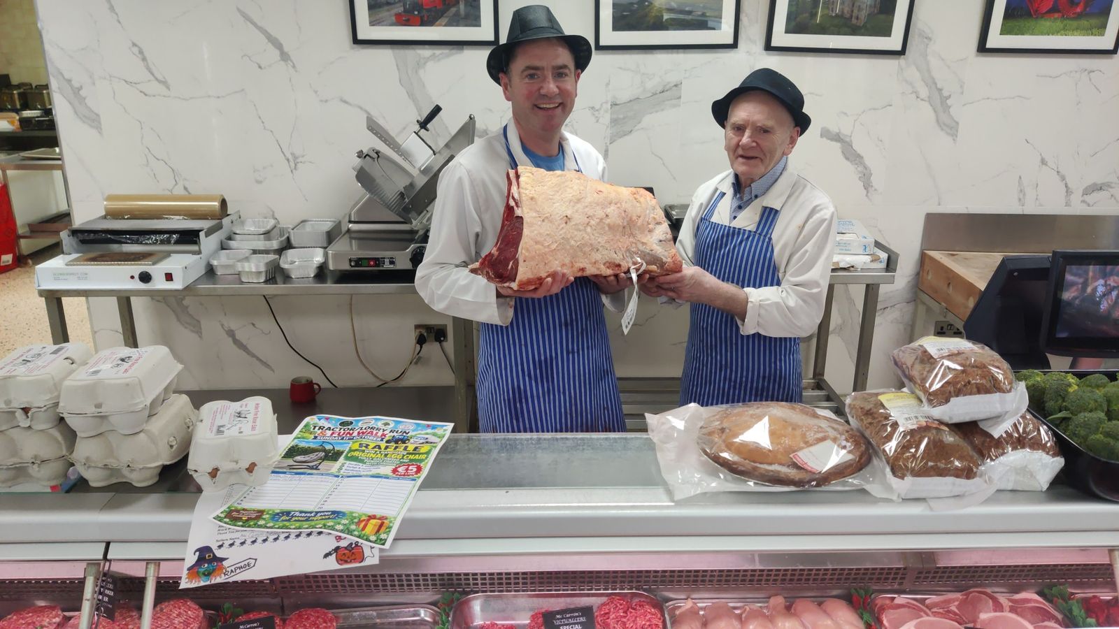 Two McCarron's butchers in trilby hats and striped aprons holding a large rib of beef behind the counter